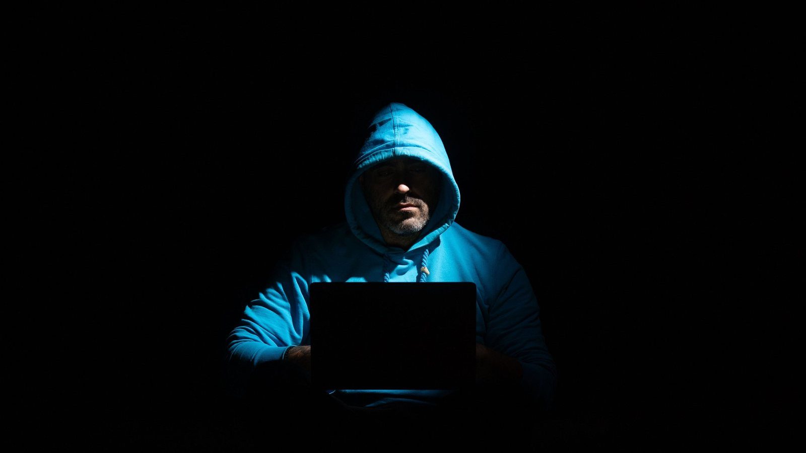A hooded figure lit only by a laptop screen in a dark room